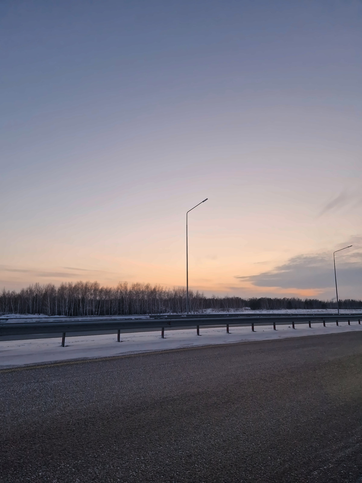 A snowy highway at dusk, a guardrail and two street lamps against an apricot horizon