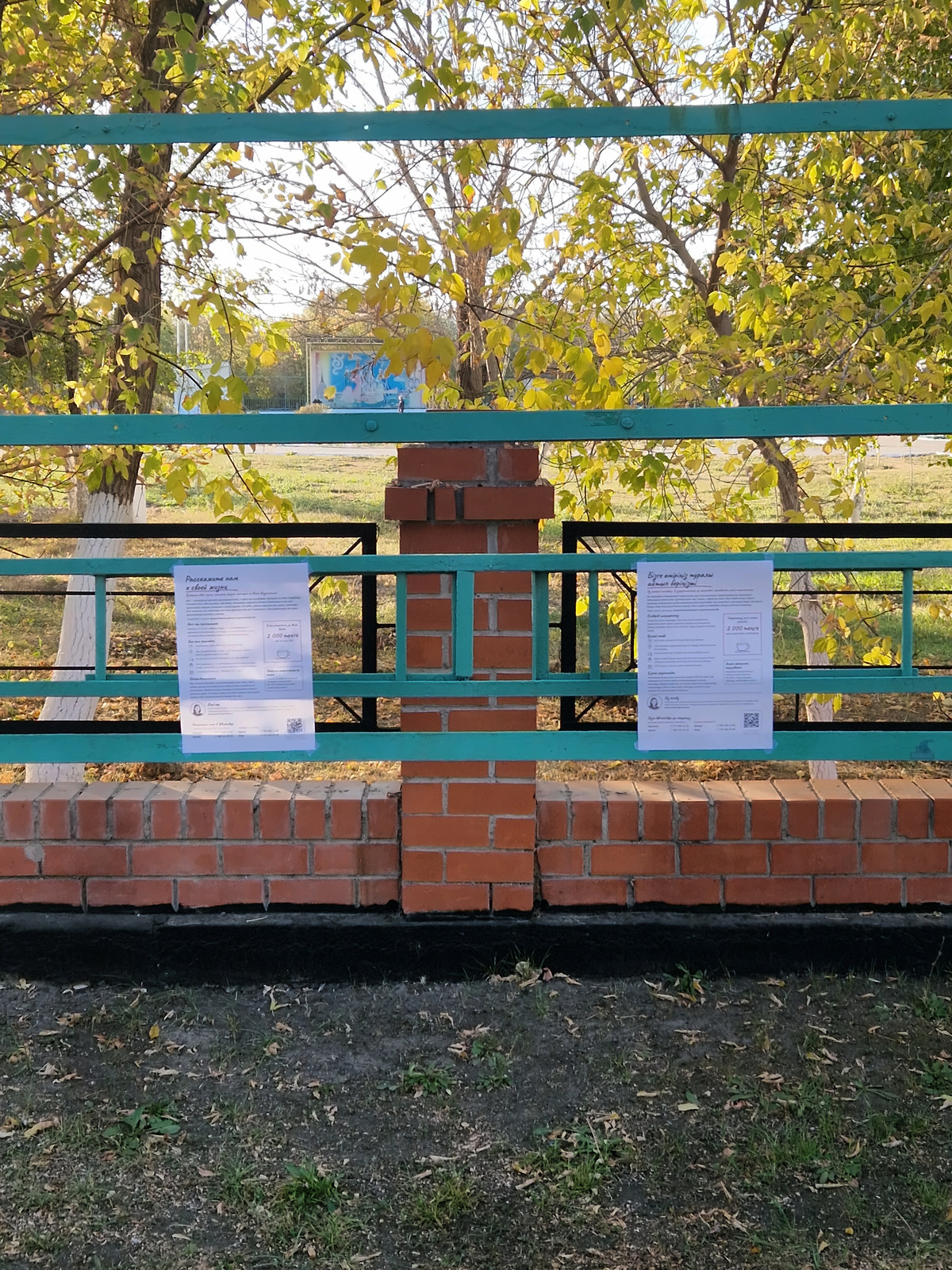 Two posters tied to a green fence under autumn trees